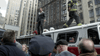 A man stands atop a fire truck on a city street, surrounded by tall buildings and crowds—an intense moment reminiscent of scenes from "Sharknado 2: The Second One (2014)" DVD.