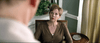 A woman with short hair in a brown cardigan sits seriously in a chair, surrounded by plants and decor, resembling a scene from the "Georgetown (2019) DVD.