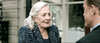 An elderly woman with long gray hair and a blue patterned jacket smiles while talking to a man outdoors, large windows behind them—perhaps reminiscing about watching the Georgetown (2019) DVD together.