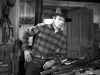 A man in a plaid shirt and cowboy hat stands indoors by a table with rifles, looking alert and ready to draw—a suspenseful scene reminiscent of classic Westerns, similar to what you’ll find in The Westerner (1940) DVD.