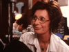 A woman with brown hair and glasses smiles in a white blouse and heart-shaped necklace. Indoors, shelves of rare movies, including the "Courage (1986) DVD," create a cozy scene with another person blurred in the background.