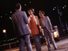 Three people in suits stand on a dimly lit street at night, car headlights casting shadows behind them. The suspenseful, cinematic scene recalls a true crime drama such as Courage (1986) DVD, with distant lights evoking classic films.