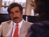 A man with dark hair and a mustache in a light suit, white shirt, and red tie sits indoors, gazing at someone with long hair—a scene reminiscent of the film "Courage (1986)" available on DVD.