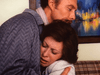 A man gently embraces a distressed woman who appears to be crying, as they sit on a plaid couch—a moving scene reminiscent of true crime dramas like Courage (1986) DVD. A colorful painting hangs on the wall behind them.