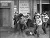 In West of the Pecos (1945) DVD, men in cowboy hats rush from the Brazos Bar as one lies on the ground; others react with drawn guns near a Mrs. Healy Dressmaker sign in this classic Western scene.