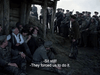 A group sits outside a wooden shelter under armed guard; one soldier stands with a rifle. Subtitles: "-Sit still! -They forced us to do it." A gripping scene from Come and See (1995) DVD.