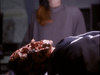 A gloved hand holds a bloody, mangled object in the foreground, evoking sci-fi horror scenes like those in Dead Space (1991); in the background, a person wearing a gray turtleneck stands blurred.