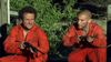 Two men in orange prison uniforms crouch outdoors with rifles, tense and alert amid green foliage—a suspenseful scene evocative of creature feature horror as seen on the Razortooth (2006) DVD.