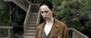 A woman with dark hair pulled back and a serious look evokes psychological horror, much like scenes from "The Night House (2020)" DVD. She wears a brown coat over gray; blurred wooden stairs and greenery form the background.