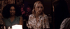 Two women sit at a dimly lit bar table with wine glasses and a candle, discussing "The Night House (2020) DVD" with a third, unseen person. In the background, other patrons enjoy themselves.