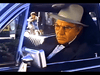 An older man sits seriously in a black car’s driver seat, glasses and light hat on—evoking the suspenseful drama of "Murder in Texas (1981)" DVD, with people and buildings seen through the window.