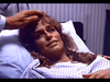 A woman with light brown hair lies in a hospital bed wearing a gown, smiling faintly as someone gently touches her forehead—a moment reminiscent of scenes from the "Murder in Texas (1981) DVD.