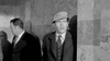 Two men in suits stand by a marble wall—one turned away, the other, in a flat cap and overcoat, looks ahead with concern. This black-and-white image evokes crime drama, fitting for "The Rise and Fall of Legs Diamond (1960) DVD.