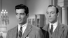 Two men in suits stand indoors, one smirking, both looking aside in a formal vintage scene—an ideal image for fans of classic crime films like The Rise and Fall of Legs Diamond (1960) DVD.