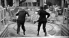Two suited men dance energetically in an empty, ornate room filled with chairs and tables. Captured in black and white, this scene recalls the vintage charm of classic films like "The Rise and Fall of Legs Diamond (1960) DVD.