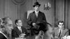 A man in a dark coat and hat stands in a formal room as others sit around a table, echoing the crime drama mood of "The Rise and Fall of Legs Diamond (1960) DVD," with elegant wood paneling and sconces enhancing the cinematic atmosphere.