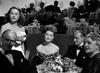 A glamorous woman holds a clutch by an elegant dining table, amid formally dressed guests at a stylish evening gathering—capturing the intrigue of classic film noir like Nightmare Alley (1947) DVD.