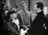 In a scene from the Virginia City (1940) DVD, a woman in a ruffled dress and fancy hat holds hands with a man in a suit as another man in a double-breasted military uniform watches, set against shelves and a map.