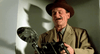 A man in a brown hat and trench coat stands indoors, smirking with a cigar and holding a vintage camera—evoking Joe Pesci’s iconic character from The Public Eye DVD (1992)—as his dramatic shadow falls on the wall behind him.
