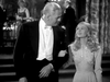 A man in a tuxedo and a woman in an embellished gown walk arm-in-arm at a formal event, evoking the elegance of "The Glass Key DVD (1942)," smiling at each other with other formally dressed guests in the background.