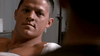 A man with short hair and a serious look sits in a room, sporting a white bandage on his shoulder and a dog tag necklace, reminiscent of a war drama. Scene from Rain (2006) DVD; the background is blurred.