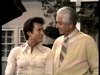Two men stand outside by a house, smiling. One has dark hair and a button-down shirt; the other, in a sweater and tie, looks like he could star in "Father Knows Best Reunion (1977)" on DVD.