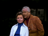 An older woman with short brown hair, in a blue turtleneck and striped shirt, sits with her eyes closed as an older man in a brown jacket stands beside her—echoing scenes from the "Father Knows Best Reunion (1977) DVD.