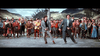Two men in suits dance energetically before a lively crowd in Scottish attire, with hills and thatched cottages behind—the scene recalls the classic film "Brigadoon (1954)" available on DVD.