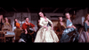 Several women in colorful 19th-century dresses dance joyfully indoors, led by a smiling woman in white—evoking classic scenes from the "Brigadoon (1954) DVD.