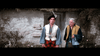 Two men in Scottish attire stand by a stone cottage, evoking classic scenes from Brigadoon (1954) DVD—one in a black beret with feather, blue shirt, white vest, and red kilt; the other with gray hair, yellow-brown jacket, and green kilt.