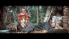 A woman in a brown dress sits on hay beside a man in gray, both smiling outdoors near trees and rustic stones—a classic scene reminiscent of Brigadoon (1954) on DVD, with wooden fencing and a barrel in the background.