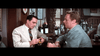 Two men in suits sit at a bar; one peels a lemon for his drink while the other listens. Bottles and framed pictures, including a Brigadoon (1954) DVD, decorate the background—hinting at rare classic films.