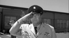 A man in uniform salutes outdoors, resembling Steve McQueen in "Soldier in the Rain (1963)" DVD. He wears a hat and medals, with a building featuring large windows visible behind him.
