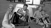 Four people in vintage outfits relax on the grass beside a golf cart, smiling and holding drinks—reminiscent of a scene from classic films like Soldier in the Rain (1963) on DVD.