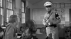 A uniformed military police officer stands holding papers, with another man seated at a desk and a prominent fan in the foreground—evoking the classic film "Soldier in the Rain (1963)" now available on DVD. Maps and documents line the office walls.