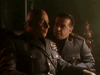 Two men in military uniforms sit close in a dimly lit room, one serious and decorated with medals. The intense scene is from the World War II drama "Countdown to War (1989) DVD.