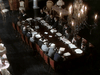 A group in formal wear sits around a grand table under chandeliers, holding a meeting reminiscent of WWII dramas like Countdown to War (1989) DVD, with documents spread out before them.