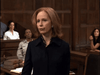 A woman with shoulder-length red hair in a dark blazer stands in a courtroom, as others watch from behind wooden walls and a table stacked with DVDs—including "Obsessed (2002)"—evoking the intensity of true-crime drama.