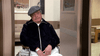 An older man in a dark jacket and flat cap sits in a wheelchair at a care facility, looking contemplative. On his lap is the "Nothing in Common (1986) DVD." The background features beige walls and a couch.