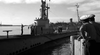 Black and white photo from "Hellcats of the Navy (1957)" DVD shows WWII submarine crew on deck at the pier, with other ships and naval personnel in the background beneath a partly cloudy sky.