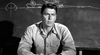 A man in a military-style shirt sits at a desk before a chalkboard with technical drawings, recalling intense WWII submarine scenes like those in the "Hellcats of the Navy (1957) DVD," as he listens or speaks intently, hands resting on the desk.