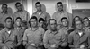 A group of twelve men in uniform shirts gather indoors with serious expressions, evoking a scene from "Hellcats of the Navy (1957)"—now available on DVD.