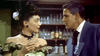 A woman in an off-the-shoulder dress and feathered hat, glass in hand, eyes a suited man with a cravat in a bottle-lined western saloon, echoing scenes from "The Bounty Hunter (1954)" DVD.