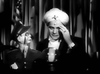 A man dressed as a fortune teller with a turban sits beside a ventriloquist dummy in uniform, evoking the classic charm of "Stage Door Canteen (1943) DVD," with a crystal ball on the table and curtains in the background.