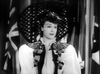 A woman in a large, lattice-patterned hat and floral jacket stands before flags in this black-and-white photo recalling wartime musical scenes from "Stage Door Canteen (1943) DVD.