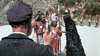 A man in a dark coat and cap faces indigenous people with spears on a rocky island—a scene reminiscent of rare finds like the Island of the Blue Dolphins (1964) DVD, with more people gathered in the background.