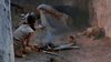 A person in primitive attire tends a smoky firepit, with pottery and a dog nearby—echoing scenes from the rare Island of the Blue Dolphins (1964) DVD.