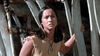 A young woman with long dark hair in a fringed tan dress stands outdoors by pale wooden poles, wearing a bold pendant—evoking scenes from Island of the Blue Dolphins (1964) DVD—as she gazes ahead with a serious look.