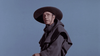 A man in a large black hat and dark cloak stands under a clear blue sky, gazing upward with a serious look—reminiscent of characters in classic films like Island of the Blue Dolphins (1964) on DVD.