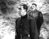 A man in a dark military uniform stands in the foreground, another behind him against a textured wall, recalling intense scenes from classic wartime dramas like "Guerrilla Girl (1953)" on DVD.